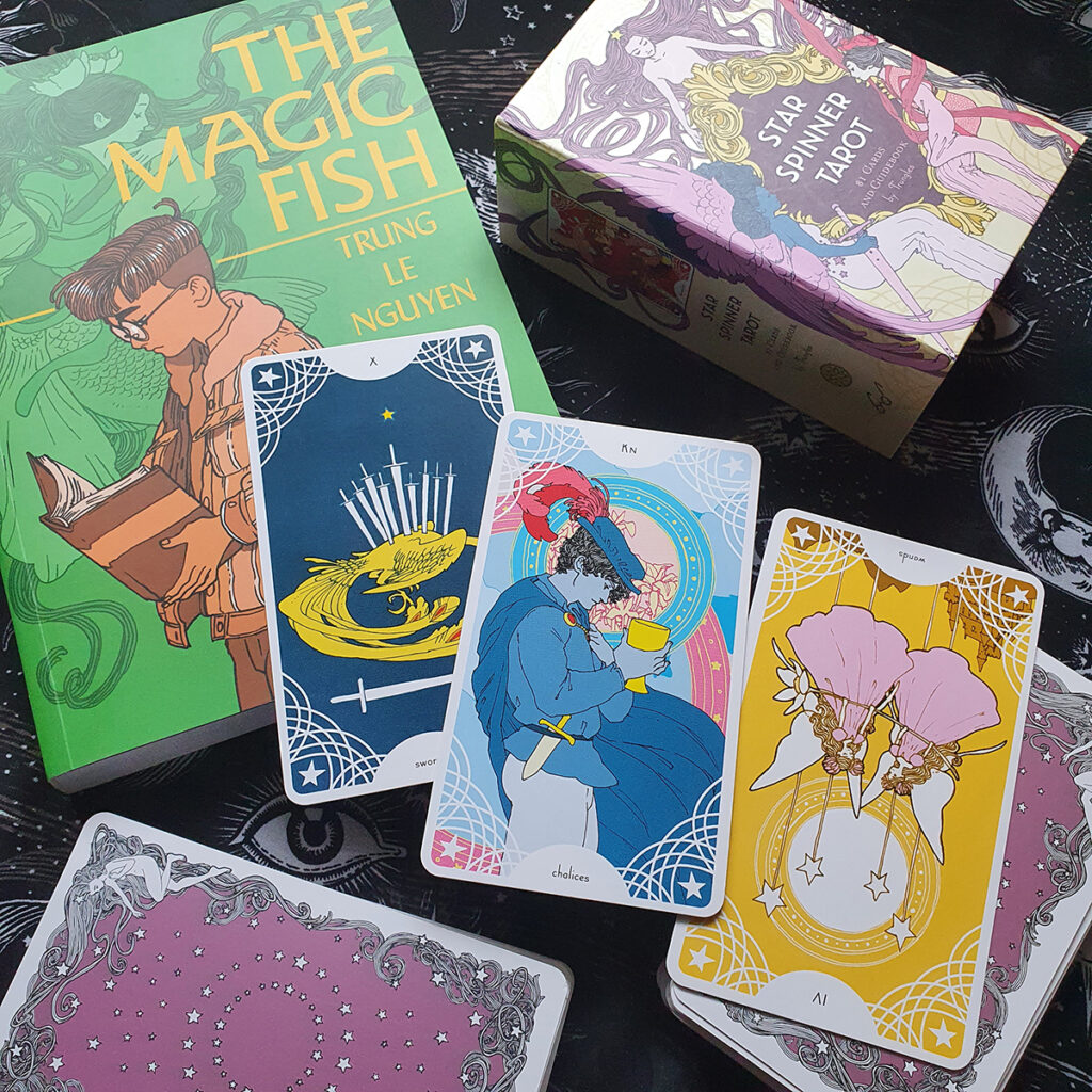 Photo of The Magic Fish graphic novel by Trung Le Nguyen and the Star Spinner Tarot box and cards, X of Swords, Knight of Chalices, VI of Wands